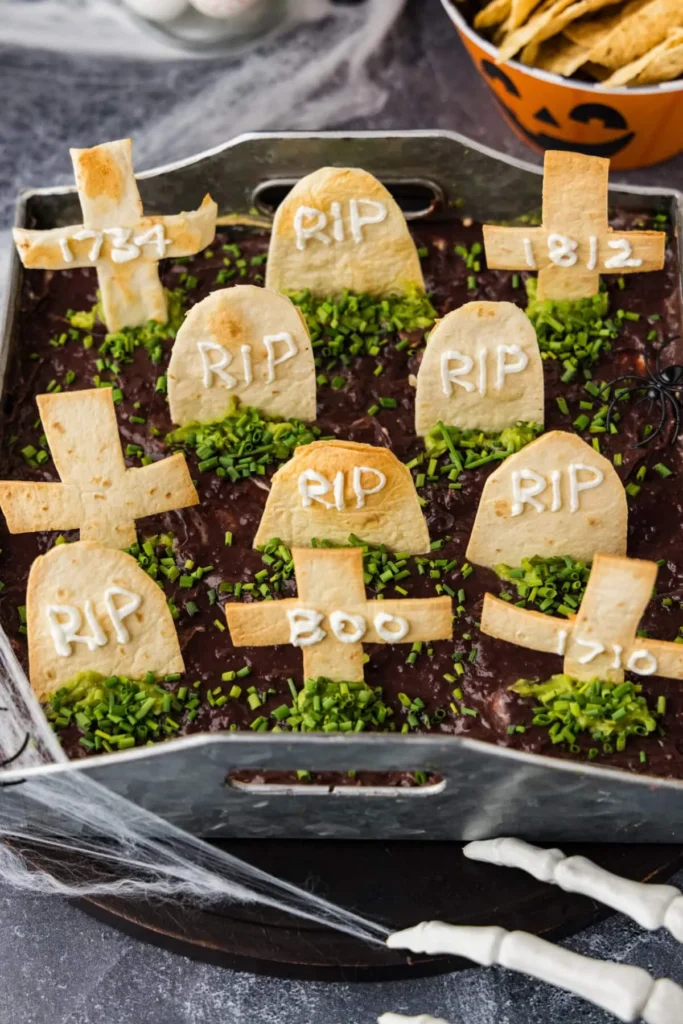 Baking dish of taco bake topped with crushed chip dirt, sour cream tombstones, and a plastic skeleton hand