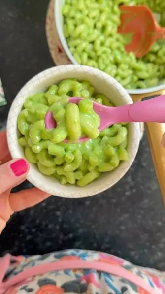Bowl of bright green macaroni and cheese with olive slice eyes and a hot sauce drizzle