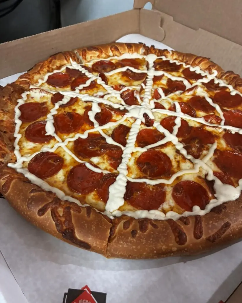 round pepperoni pizza with a white sour cream spiderweb and a black olive spider in the center